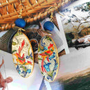 Σκουλαρίκια γυναικεία κρεμαστά με μπλε χάντρα και οβάλ στολίδι με λουλούδια και ερωδιό.